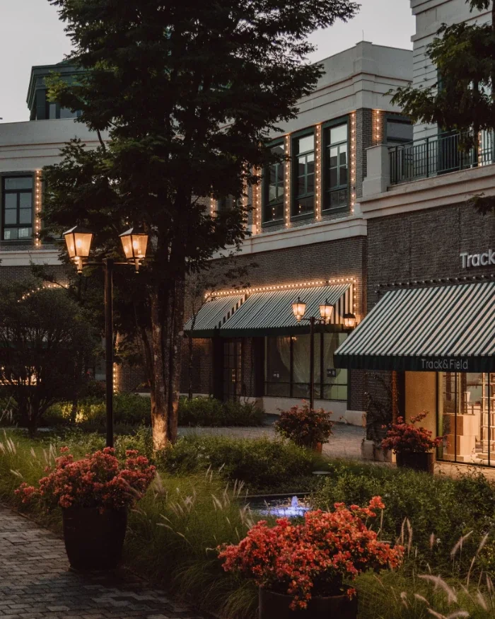 Fachadas comerciais e área de convivência do CJ Boa Vista Village, em Porto Feliz, com lojas, paisagismo e iluminação ao entardecer.
